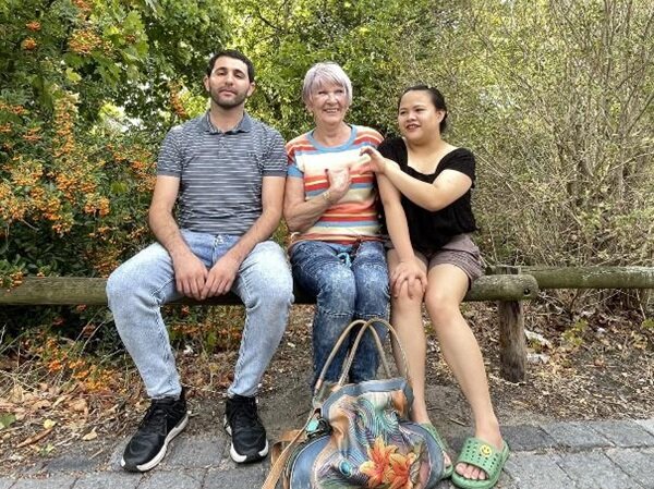 Zwei Teilnehmende sitzen mit ihrer Dozentin auf einem Holzbalken und posieren für ein Abschiedsfoto. 