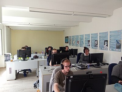 Bericht zur Berufsschulschließung am 08.05.2025 Ein heller, moderner Schulungsraum mit mehreren Reihen an Tischen, ausgestattet mit Computern und Monitoren. Junge Erwachsene sitzen mit Kopfhörern vor den Bildschirmen und konzentrieren sich auf ihre Aufgaben. An der Wand hängen informative Plakate.