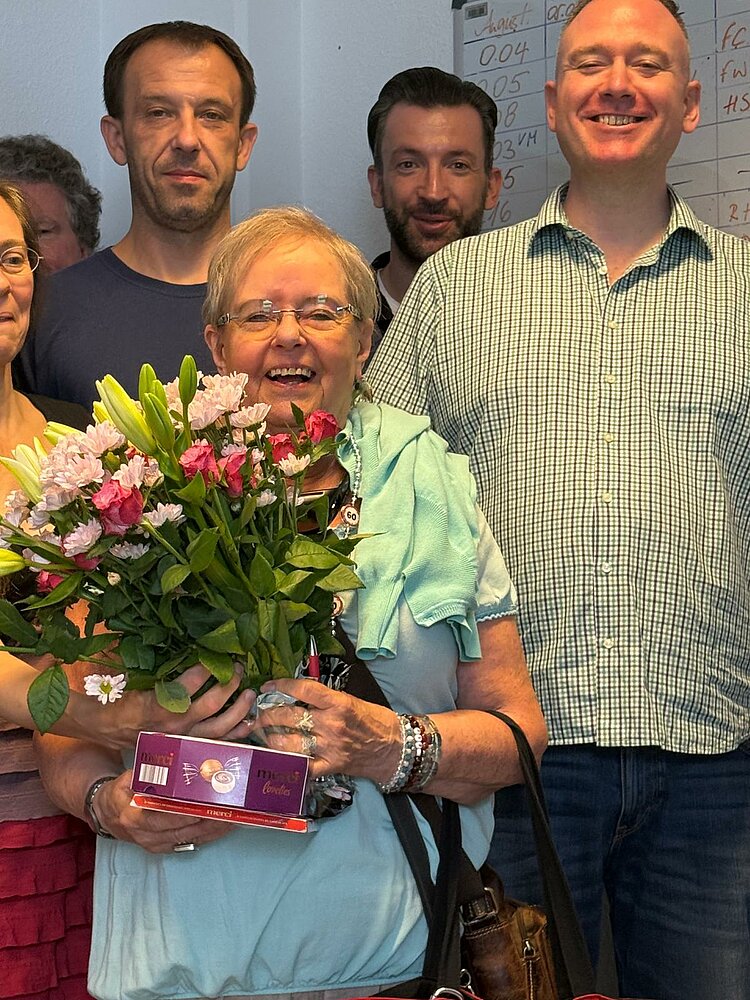 Bruni Drewitz – Die Geheimwaffe der Erwachsenenbildung Nahaufnahme einer älteren Frau, die einen Blumenstrauß und eine Schachtel Pralinen hält. Sie steht lächelnd vor mehreren Personen, die sie umgeben. Im Hintergrund ist ein beschriftetes Whiteboard sichtbar.