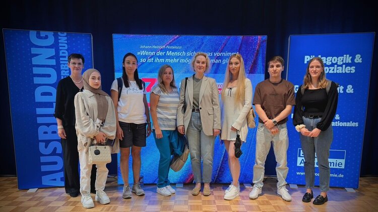 Vor einer blauen Wand mit digitalen Roll-ups der Euro Akademie steht in einem Raum eine Gruppe jüngerer und älterer Frauen sowie ein junger Mann. 