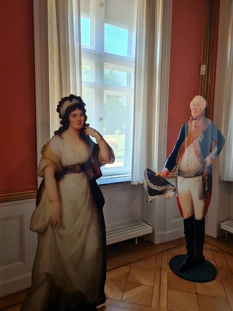 Toller Teamausflug ins Marmorpalais Zwei Figuren aus Pappe, die wahrscheinlich die ehemaligen adeligen Bewohner des Schlosses darstellen sind in einem Raum aufgestellt.