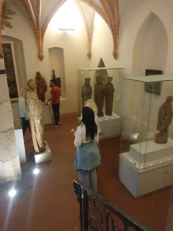 Eine Gruppe von Menschen betrachtet Ausstellungsstücke in einem Museum. Die Exponate sind hinter Glasvitrinen ausgestellt und umfassen historische Artefakte und Skulpturen. Die Besucher und Besucherinnen sind in einer hellen, architektonisch interessanten Umgebung.