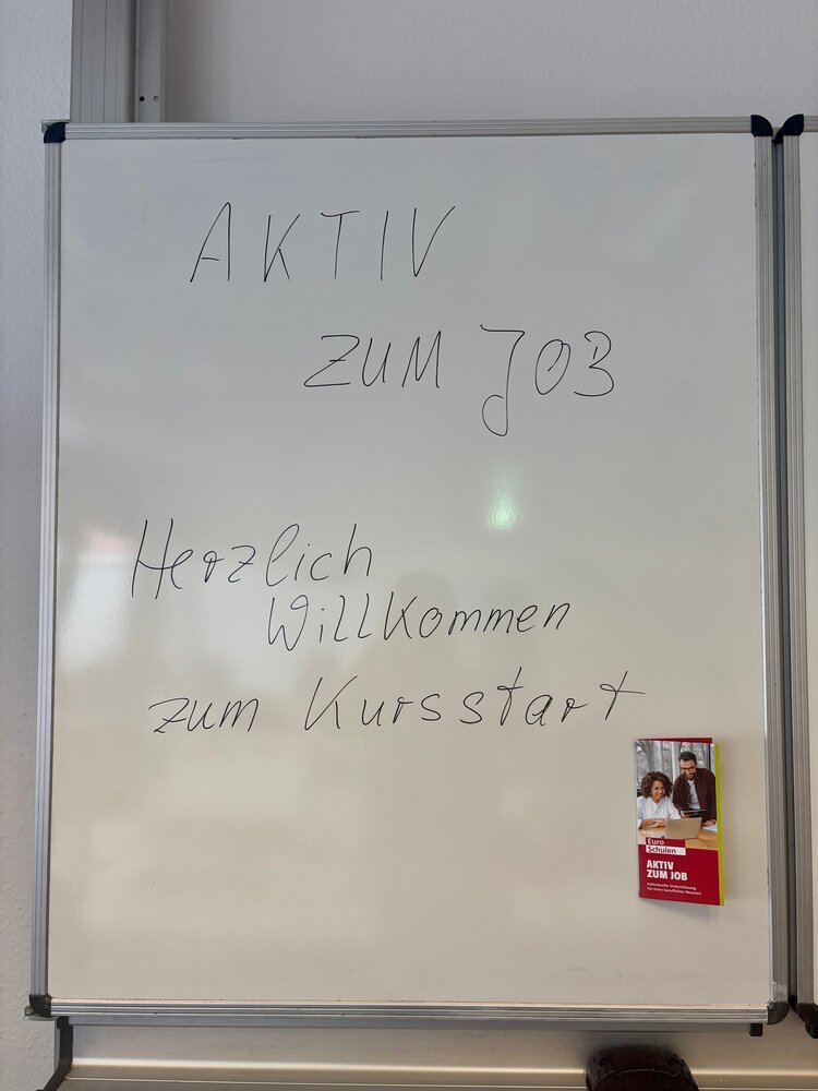 Auf einem Whiteboard steht in schwarzer Schrift "Aktiv zum Job - Herzlich Willkommen zum Kursstart"; darunter hängt der zugehörige Flyer.