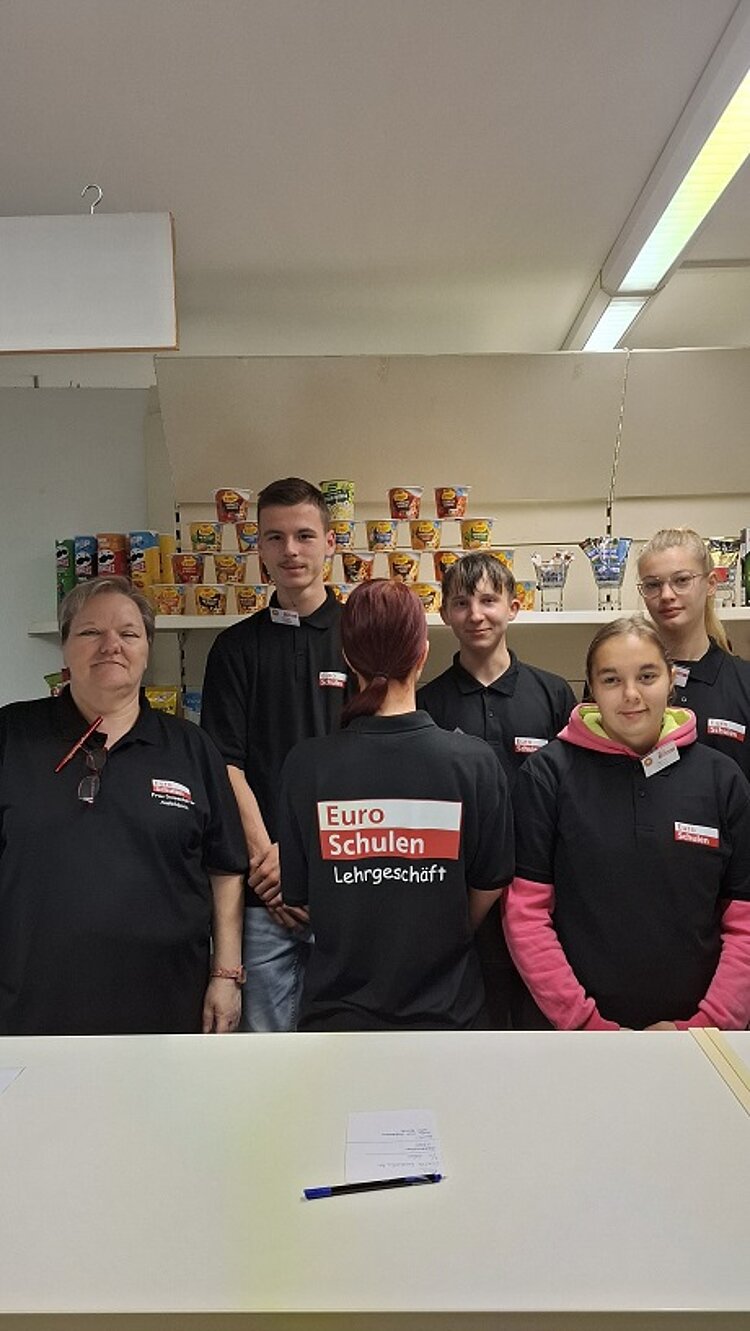 Alles neu macht der… September Drei Schülerinnen, zwei Schüler und eine Lehrerin stehen hinter der Theke des Lehrgeschäfts. Sie tragen über ihren eigentlichen Kleidungstücken Polohemden als Arbeitsschutzbekleidung. Um auch die Rückseite des Shirts zu zeigen, hat sich eine der Schülerinnen umgedreht. In den Regalen hinter den Personen stehen Produkte, die man aus dem Supermarkt kennt, darunter Chips und Fertigsuppen.