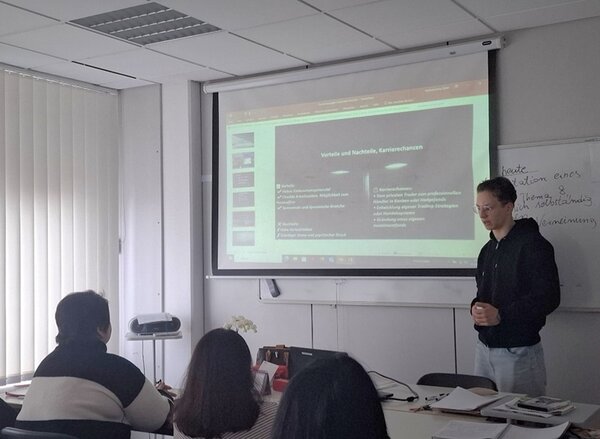 Ein männlicher Teilnehmender steht vorn am Whiteboard, er trägt einen dunklen Kapuzenpullover und seine Präsentation vor.
