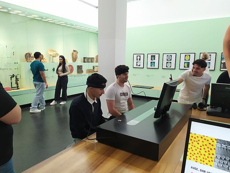 Sonniger Exkursionstag der Euro-Schulen Dresden: Spannende Einblicke in Wissenschaft und Kultur In einem interaktiven Museum sitzen zwei junge Männer mit Stirnbändern vor einem Bildschirm, während eine weitere Person lachend auf das Display zeigt.