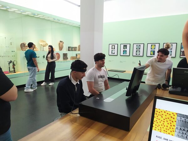 In einem interaktiven Museum sitzen zwei junge Männer mit Stirnbändern vor einem Bildschirm, während eine weitere Person lachend auf das Display zeigt.