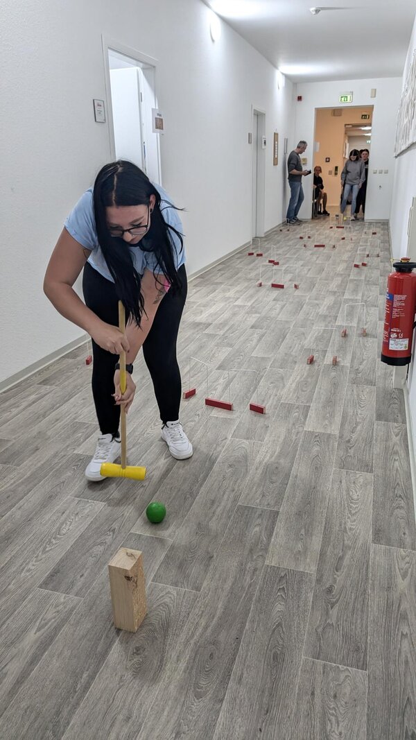 Mini-Cricket im Flur. Eine Schülerin versuch eine grüne Kugel durch aus mit roten Holzbausteinen gebaute Tore zu manövrieren.