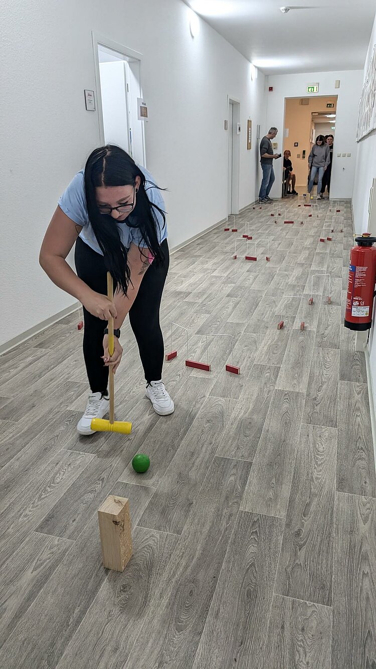 Rückblick Sportfest 2024 Mini-Cricket im Flur. Eine Schülerin versuch eine grüne Kugel durch aus mit roten Holzbausteinen gebaute Tore zu manövrieren.