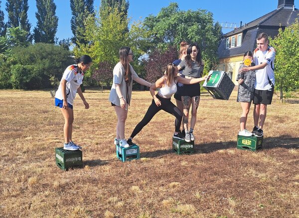 Mehrere Schülerinnen und Schüler stehen auf vier umgedrehten Bierkästen, teilweise befinden sich zwei Personen zusammen auf einem Kasten. Eine Schülerin wechselt gerade von einem Kasten zum nächsten und übergibt gerade einen Bierkasten an ein Teammitglied auf dem letzten Kasten.