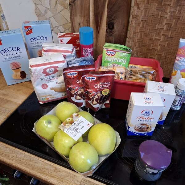 Unterschiedliche Backzutaten, darunter Zucker, Mehl, Glasur, Äpfel, Puderzucker, Backpulver, Streusel