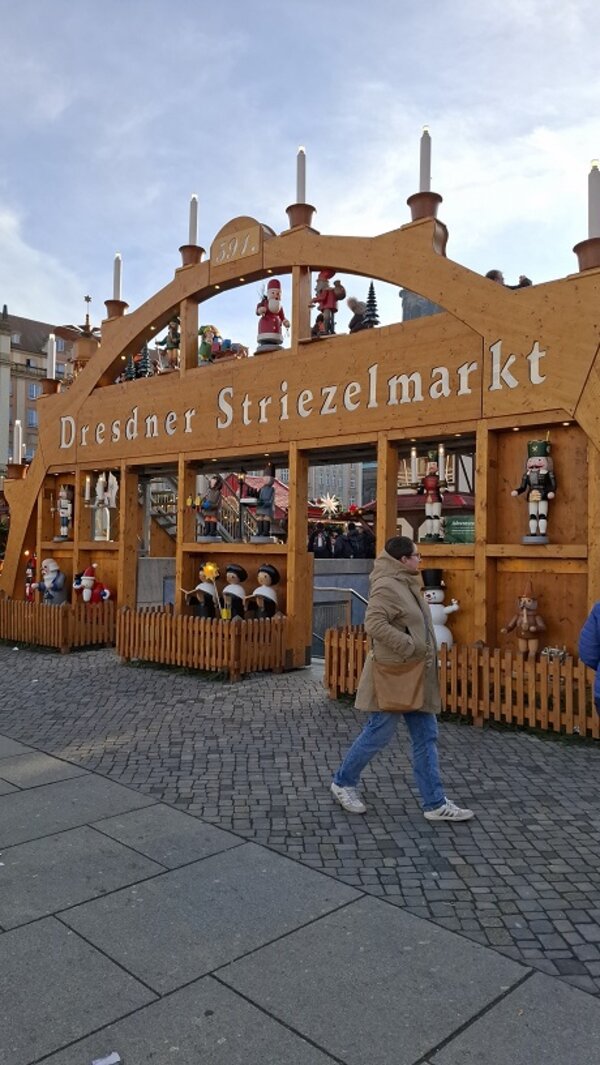 Holzportal mit der Aufschrift ‚Dresdner Striezelmarkt‘, dekoriert mit Figuren und Kerzen, davor gehen Menschen über den Platz