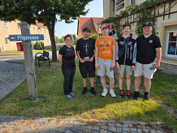 Die vier männlichen Auszubildenden zu Fachlageristen posieren, gemeinsam mit ihrer Lehrerin, für ein Gruppenfoto an einem Schild, auf dem Pilgeroase steht. 
