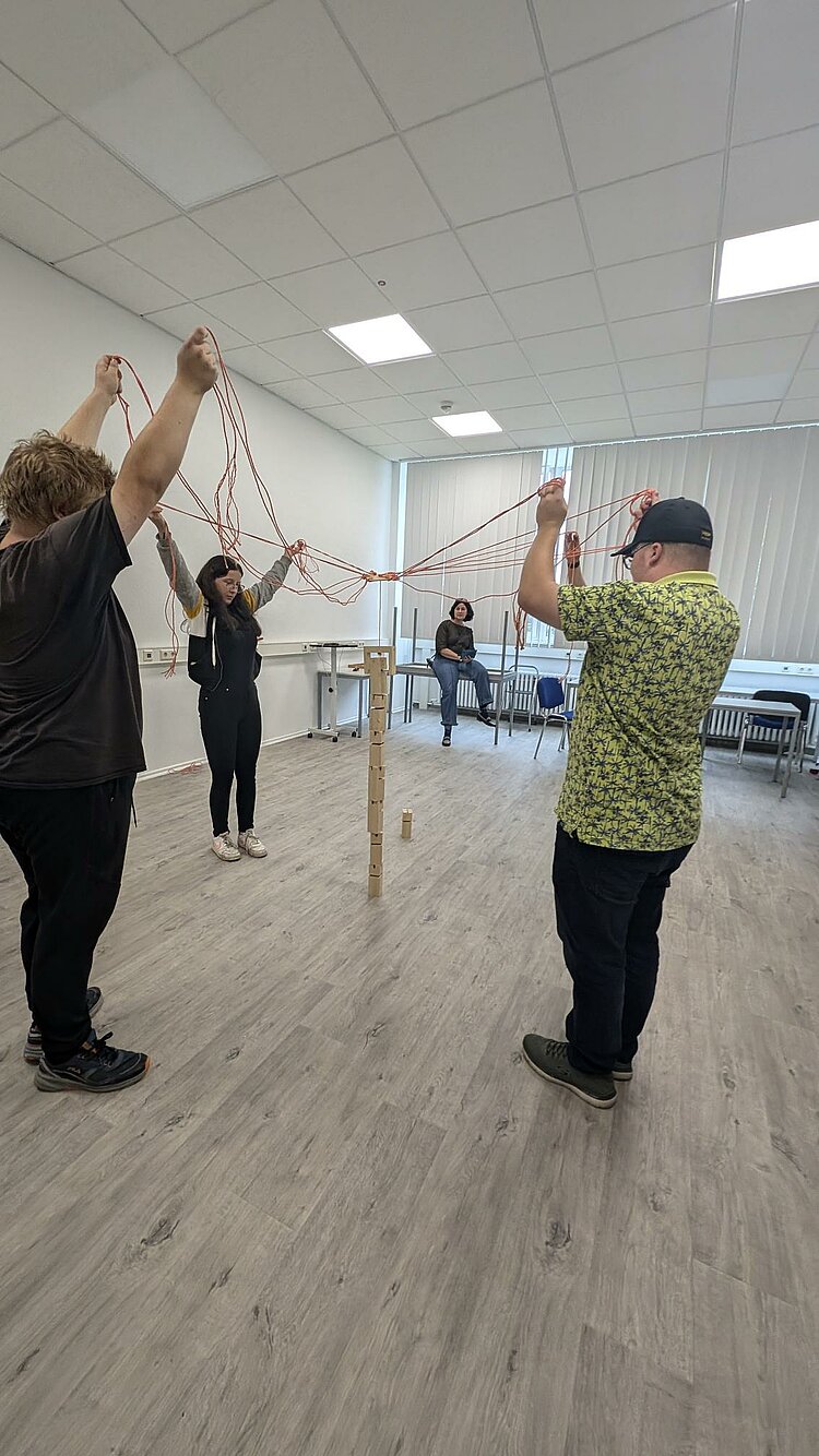 Rückblick Sportfest 2024 Geschicklichkeitsspiel. Schüler versuchen mit verschiedenen Leinen aus Kordel und einer Öffnung am Schnittpunkt Holzbausteine zu umspannen, diese anzuheben und damit einen Turm zu bauen.