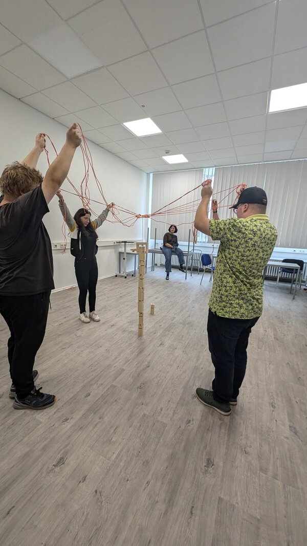 Geschicklichkeitsspiel. Schüler versuchen mit verschiedenen Leinen aus Kordel und einer Öffnung am Schnittpunkt Holzbausteine zu umspannen, diese anzuheben und damit einen Turm zu bauen.
