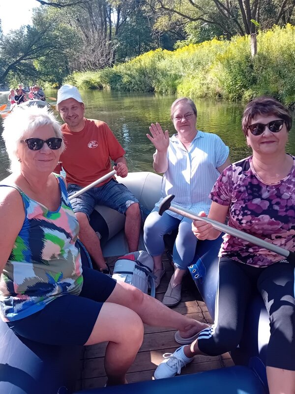 In einem blauen Schlauchboot sitzen drei Frauen und ein Mann. Zwei Frauen tragen Sonnenbrille, eine Frau winkt in die Kamera. Zwei Personen halten ein Ruder in der Hand. Im Hintergrund ist ein zweites Boot erkennbar.