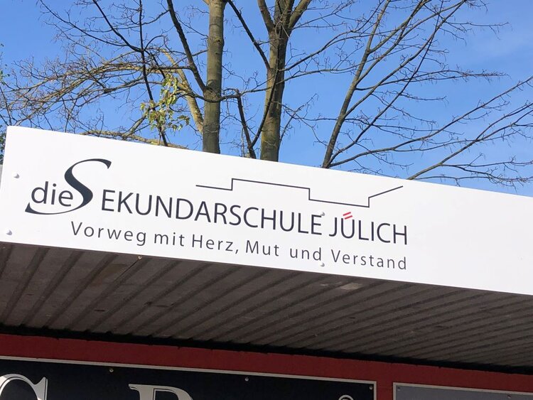 Schild am Eingang der Sekundarschule Jülich mit dem Leitspruch „Vorweg mit Herz, Mut und Verstand“ vor einem klaren blauen Himmel und kahlen Bäumen.