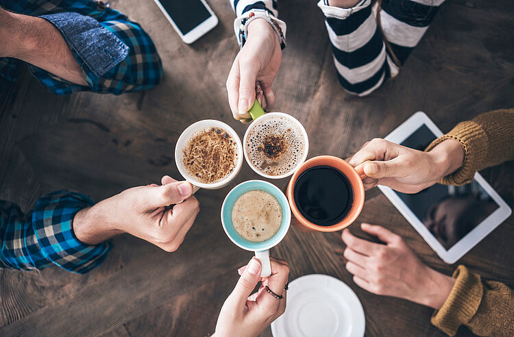 Neues Angebot: „Sprachcafé“ für Geflüchtete Der Blick ist von oben auf eine Holztisch gerichtet, an dem mehrere Hände mit Kaffeetassen anstoßen. Es handelt sich um Männer- und Frauenhände, die Tassen sind mit unterschiedlichen Kaffeekreation gefüllt. Außerdem liegen ein Tablet und ein Smartphone auf dem Tisch.