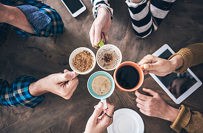 Neues Angebot: „Sprachcafé“ für Geflüchtete Der Blick ist von oben auf eine Holztisch gerichtet, an dem mehrere Hände mit Kaffeetassen anstoßen. Es handelt sich um Männer- und Frauenhände, die Tassen sind mit unterschiedlichen Kaffeekreation gefüllt. Außerdem liegen ein Tablet und ein Smartphone auf dem Tisch.