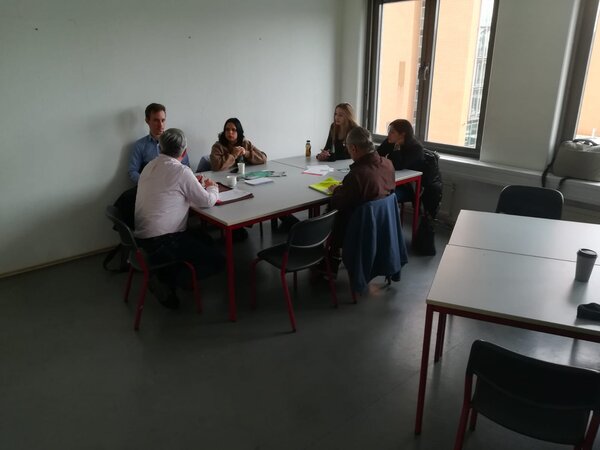 Sechs Personen sitzen an einem Tisch und besprechen Unterlagen; Notizen und Getränke liegen auf dem Tisch, Fenster rechts.