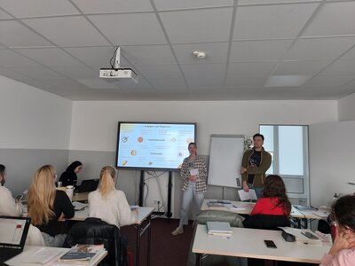 Zwei Personen stehen vor einem Bildschirm in einem Klassenzimmer und halten eine Präsentation über verschiedene Aufgaben- und Tätigkeitsbereiche. Mehrere Zuhörer sitzen an ihren Tischen und verfolgen die Präsentation.