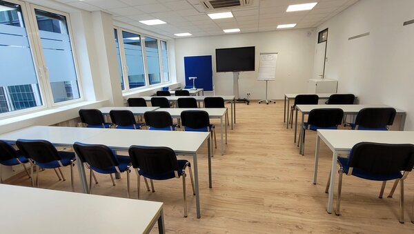Heller Unterrichtsraum mit mehreren Reihen weißer Tische und schwarzen Stühlen, vorne ein großer Bildschirm, Flipchart und Dokumentenkamera, links eine Fensterfront.