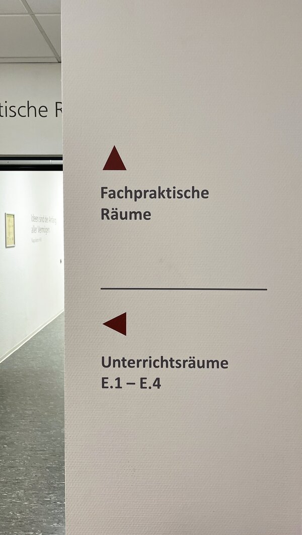 Nahaufnahme eines Wegweisers an der Wand mit Pfeilen und der Beschriftung „Fachpraktische Räume“ sowie „Unterrichtsräume E.1 – E.4“.