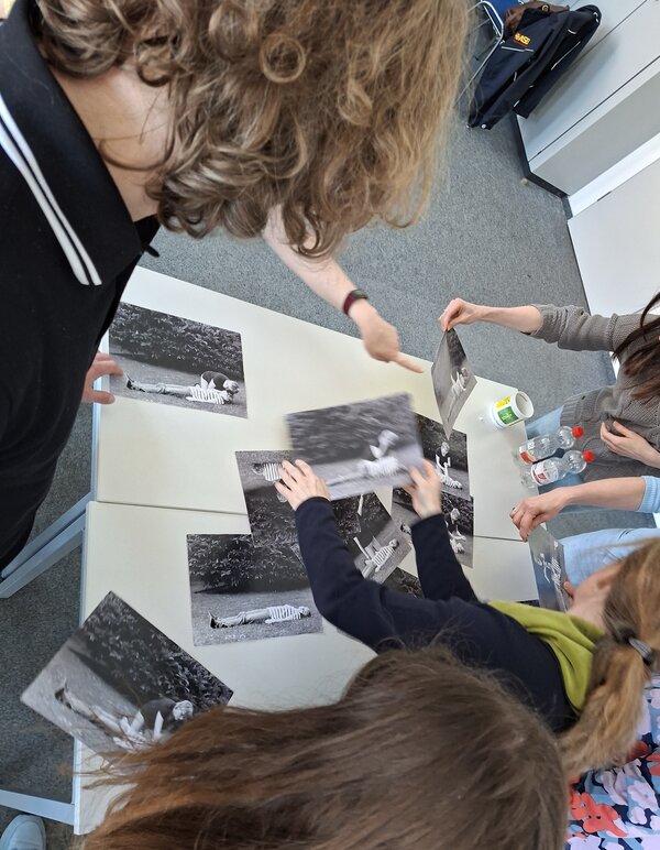 Auf einem Tisch liegen mehrere Schwarz-Weiß-Fotos; mehrere Personen stehen um den Tisch und ordnen die Fotos.