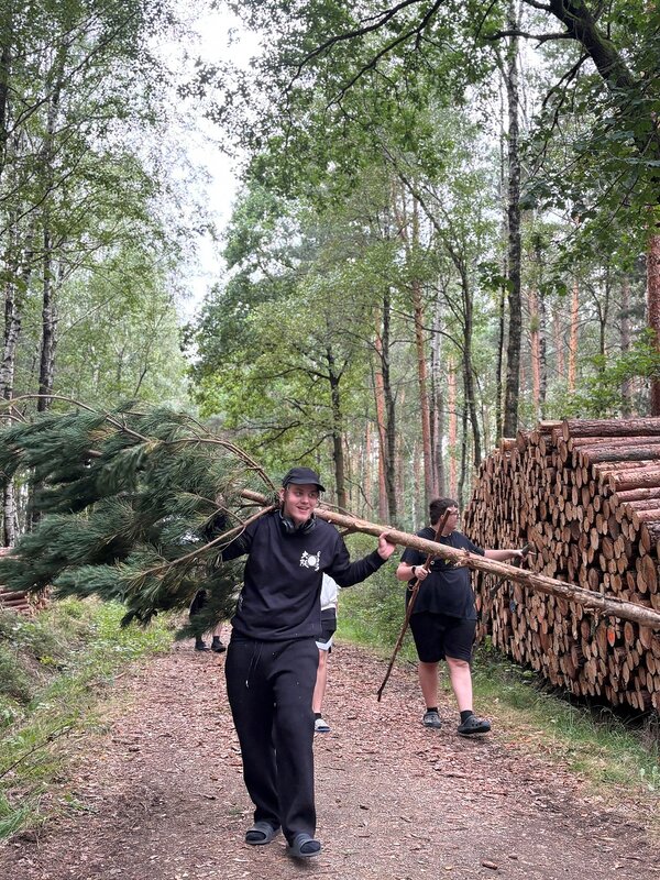 Ein junger Mann trägt einen Nadelbaum über der Schulter. Hinter ihm trägt ein weiterer junger Mann zwei lange Stöcke in den Händen. Beide gehen auf einem Waldweg. Rechts vom Weg ist ein Stapel geschnittener Baumstämme zu sehen.