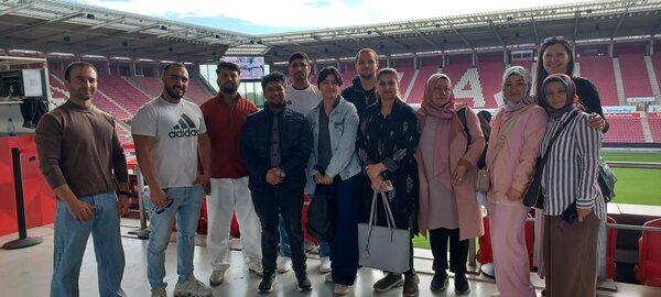 Eine Gruppe von zwölf lächelnden Personen steht zum Gruppenbild in einem Sportstadion.