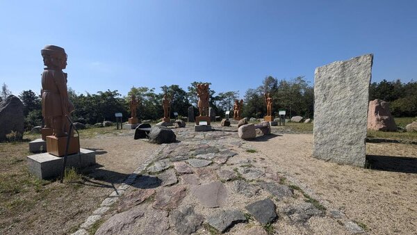 Zwischen Steinblöcken stehen überlebensgroße Holzfiguren auf einem Platz, der teilweise mit großen Steinen gepflastert ist. Im Hintergrund stehen Bäume.