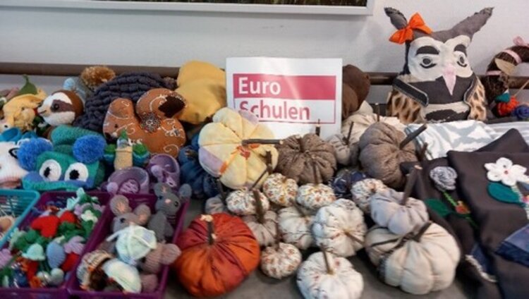 Viele handgemachte Stofffiguren, wie Kürbisse, Mäuse, Eulen sind auf einem Tisch augebreitet. In der Mitte ist ein Schild mit dem Logo der Euro-Schulen.