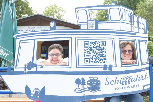 Zwei Frauen schauen durch die Fenster einer großen Fotowand in Form eines Schiffes mit der Aufschrift „Schifffahrt Hohenwarte“.