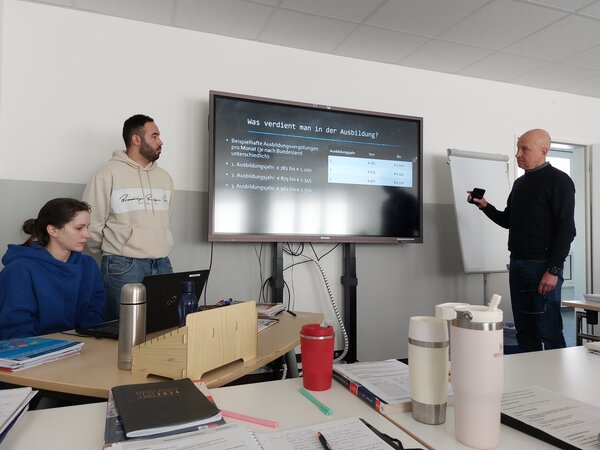 Ein Mann in dunkler Kleidung präsentiert mit einem Laserpointer eine Folie zur Ausbildungsvergütung. Neben ihm steht ein junger Mann im Kapuzenpulli. Eine Teilnehmerin sitzt an einem Laptop, während weitere Notizbücher, Trinkbecher und Schreibutensilien auf den Tischen liegen.
