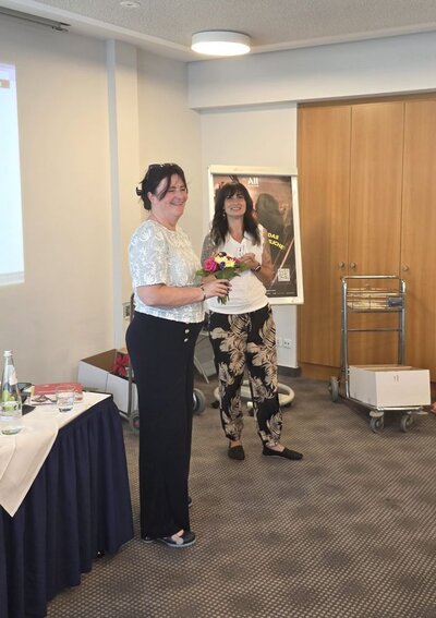 Zwei Frauen stehen lächelnd nebeneinander in einem Konferenzraum. Die Frau links trägt ein weißes, gemustertes Oberteil und eine schwarze Hose und hält einen Blumenstrauß in den Händen, während die Frau rechts ein weißes Oberteil und eine schwarz-beige gemusterte Hose trägt.