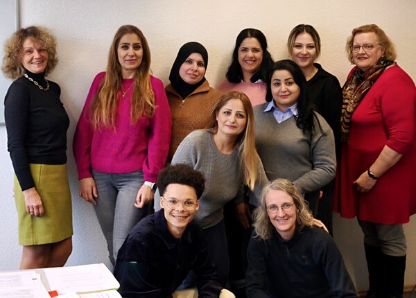 Eine Gruppe von Teilnehmenden der Euro-Schulen Hannover steht und sitzt vor einer weißen Wand und posiert für ein gemeinsames Gruppenfoto zum Kursstart der Qualifizierung. Die stehenden Personen tragen unterschiedliche Oberteile in kräftigen Farben wie Pink, Rot und Schwarz sowie verschiedene Hosen und Röcke. Eine Person trägt einen Rock in Olivgrün und einen schwarzen Pullover, eine andere ein rotes Kleid mit gemustertem Schal. Im Vordergrund sitzen zwei Personen, eine mit dunklem Oberteil und eine mit langem, hellblondem Haar. Auf einem Tisch im Vordergrund liegen einige Blätter Papier.
