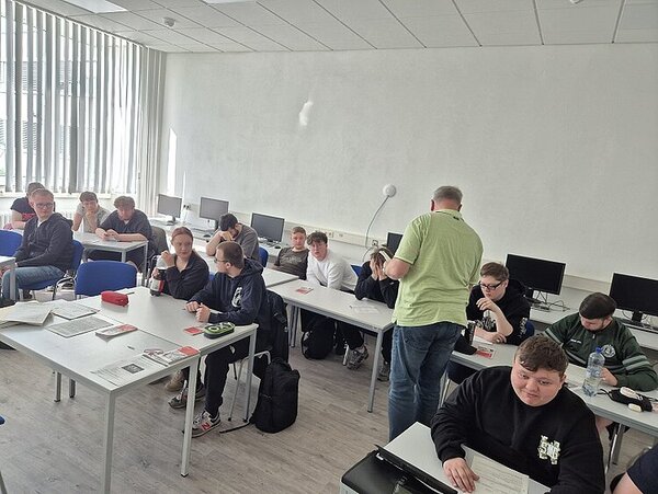 Eine Gruppe von Schülern sitzt in einem Computerraum an weißen Tischen. Einige unterhalten sich oder arbeiten mit Arbeitsblättern. Ein Lehrer in grünem Hemd steht in der Mitte des Raumes und spricht mit einer Schülergruppe. Die Stimmung wirkt konzentriert und locker.