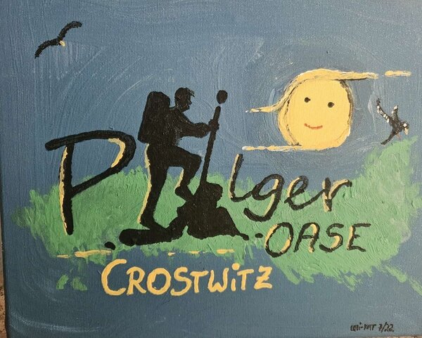 Ein handgemaltes Schild mit der Aufschrift 'Pilger-Oase Crostwitz'. Im Hintergrund sieht man eine stilisierte Figur eines Pilgers mit Wanderstock sowie eine lächelnde Sonne. Das Schild ist in blauen, grünen und gelben Farben gestaltet.
