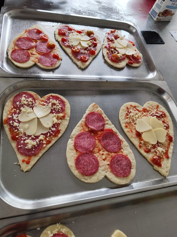 Mehrere ungebackene, herzförmige Pizzen auf einem Backblech. Sie sind mit Salami, Käse und Tomaten belegt, einige mit dekorativen Käsestücken in Blumenform.