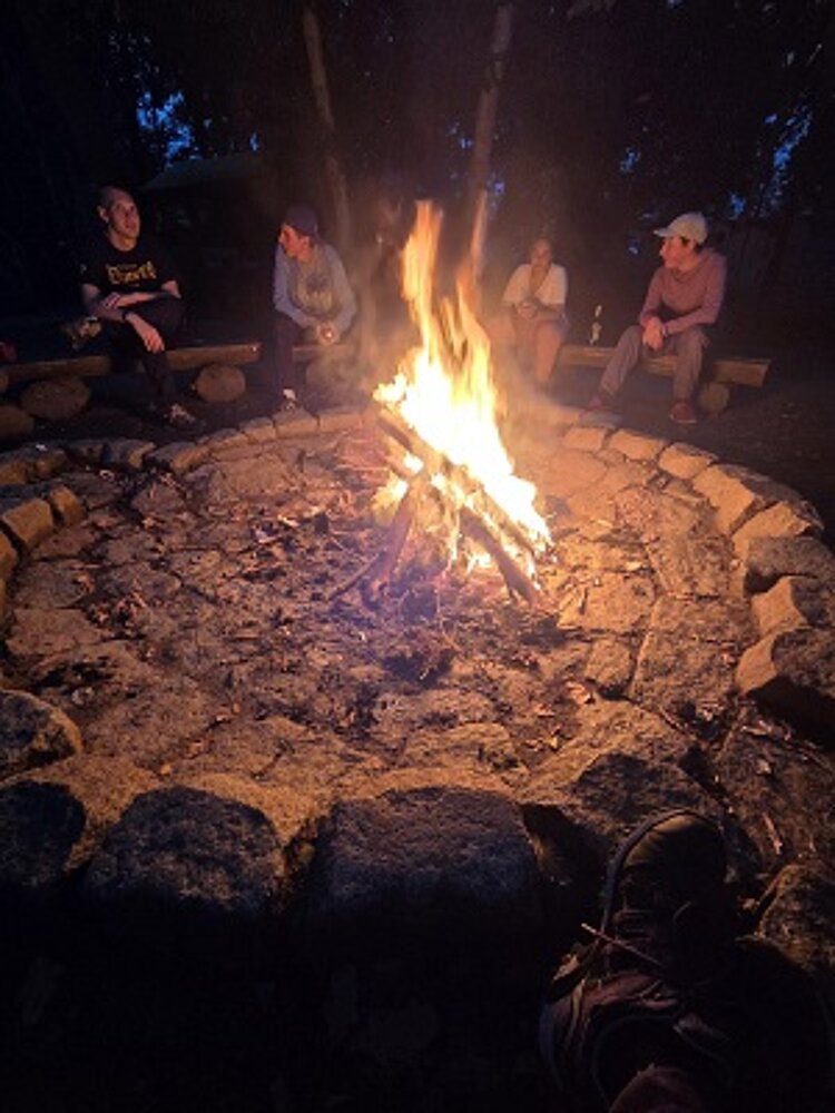 Lagerfeuer am Abend in einer steinernen Feuerstelle, mehrere Personen sitzen entspannt im Hintergrund.