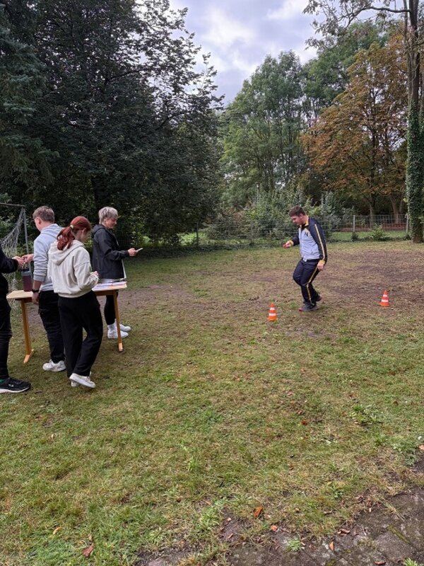 Eine Gruppe Jugendlicher steht im Freien auf einer Wiese neben einem Holztisch. Eine Person läuft oder springt zwischen zwei orangefarbenen Hütchen hindurch. Im Hintergrund sind Bäume und ein Zaun zu sehen. Die Szene wirkt wie eine Gruppenaktivität oder ein Spiel im Freien.
