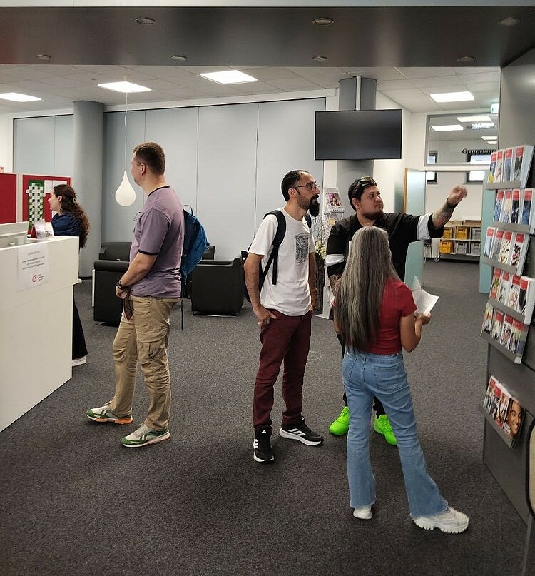 Gruppe IK-359 zu Besuch im BIZ Eine Gruppe von Teilnehmenden des Integrationskurses steht im Berufsinformationszentrum vor einem Aufsteller mit Informationsbroschüren. Ein Teilnehmer greift gerade nach einer Broschüre.