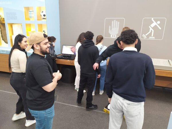 Besucher eines Museums stehen vor einer interaktiven Wand mit verschiedenen Symbolen und versuchen, ein Experiment mit einem Paar Bürstenschuhe.