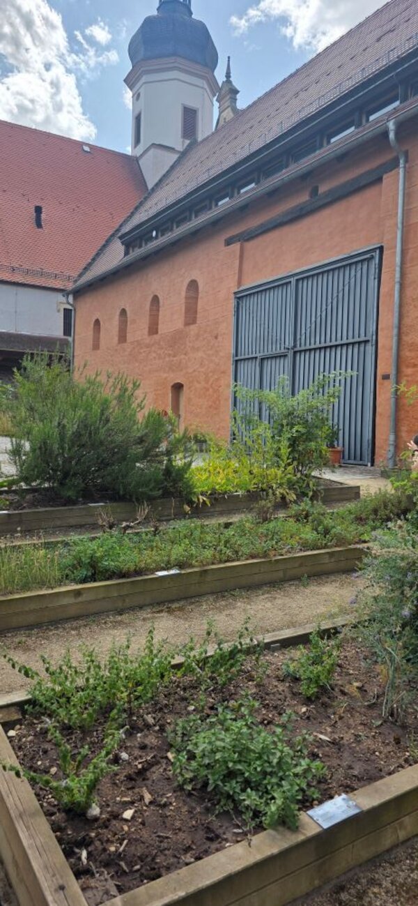 Eine restauriertes, scheunenähnliches Gebäude mit einer Fassade aus Lehm und großen grauen Stahltür. Im Vordergrund sind ein gepflegter Garten mit Kräuterbeeten und im Hintergrund ein Teil einer Kirche zu sehen. Der Himmel ist leicht bewölkt.
