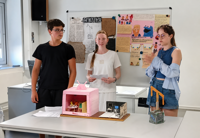 Drei Jugendliche präsentieren vor einer Pinnwand mit Plakaten zwei kleine Modellbauten auf einem Tisch. Im Hintergrund ist ein heller Klassenraum mit Fenstern und weiteren Unterrichtsmaterialien zu sehen.