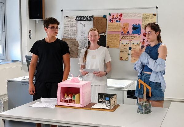 Drei Jugendliche präsentieren vor einer Pinnwand mit Plakaten zwei kleine Modellbauten auf einem Tisch. Im Hintergrund ist ein heller Klassenraum mit Fenstern und weiteren Unterrichtsmaterialien zu sehen.