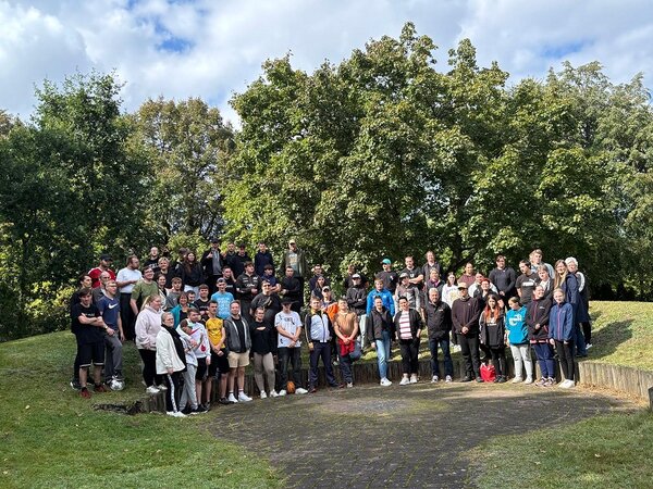 Eine große Gruppe junger Auszubildender Menschen und Erwachsener steht für ein Gruppenfoto vor einem großen, grünen Baum auf einer Wiese an einem sonnigen Tag.