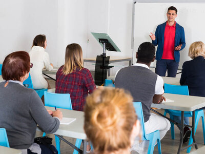 Ein Mann steht vor einem Whiteboard in einem Klassenzimmer und hält einen Vortrag. Er trägt ein rotes Hemd und eine blaue Jacke. Die Teilnehmer sitzen an Tischen mit blauen Stühlen und hören aufmerksam zu. Einige Teilnehmer machen sich Notizen. Im Hintergrund ist ein Podium mit einem Laptop zu sehen.