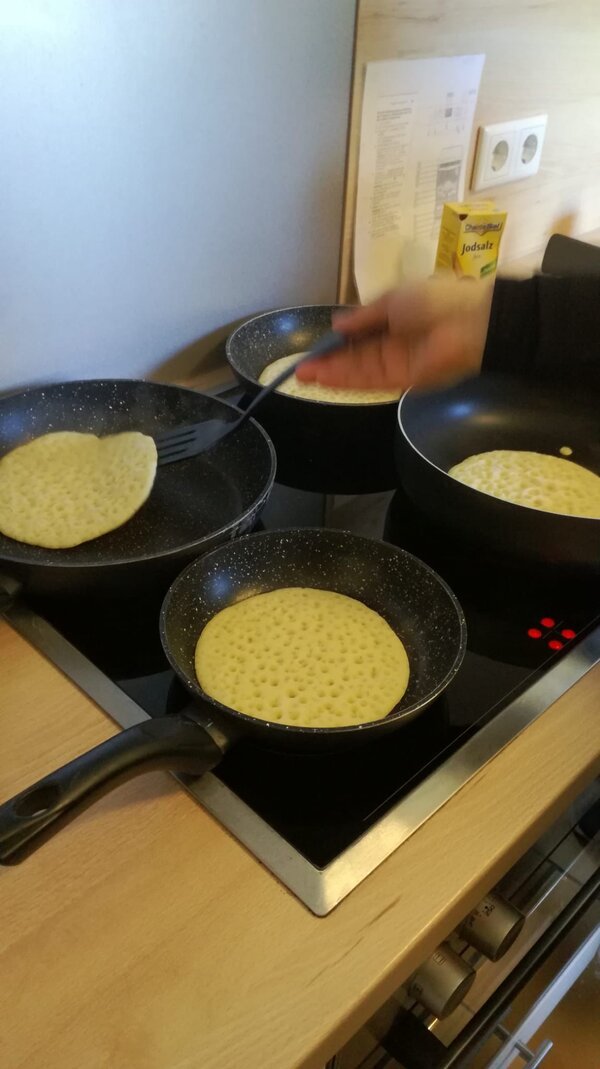 Mehrere Pfannen stehen auf einem Herd, in denen Teigfladen mit vielen kleinen Blasen gebacken werden. Eine Hand hält einen Pfannenwender und hebt einen der Fladen an.