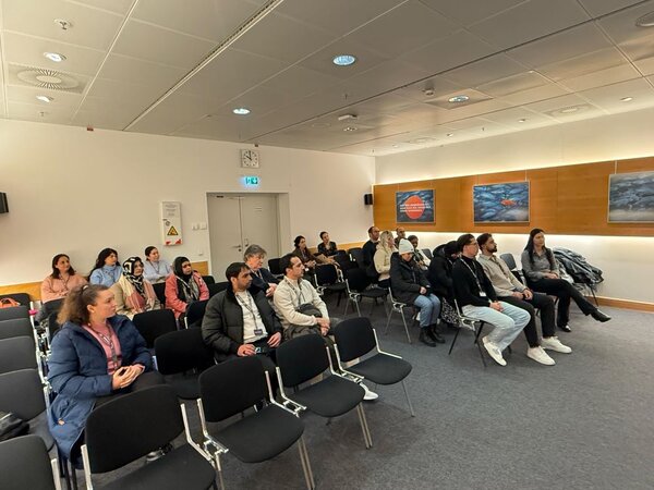 Eine Gruppe von Menschen sitzt in einem Seminarraum auf Stühlen in Reihen. Die Personen sind unterschiedlich gekleidet, einige tragen Jacken oder Pullover. Der Raum ist hell und freundlich mit großen Fenstern und einer Uhr an der Wand. Im Hintergrund sind Bilder an der Wand zu sehen. Die Atmosphäre wirkt konzentriert und aufmerksam, als ob eine Präsentation oder ein Vortrag stattfindet.